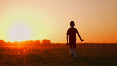 Tracking-a-boy-football-player-in-a-red-t-shirt-and-shorts-running-with-the-ball-at-sunset-in-the-field-on-the-grass.-The-concept-of-young-players.-The-young-football-player-dreams-of-a-professional-career-and-trains-in-the-field.